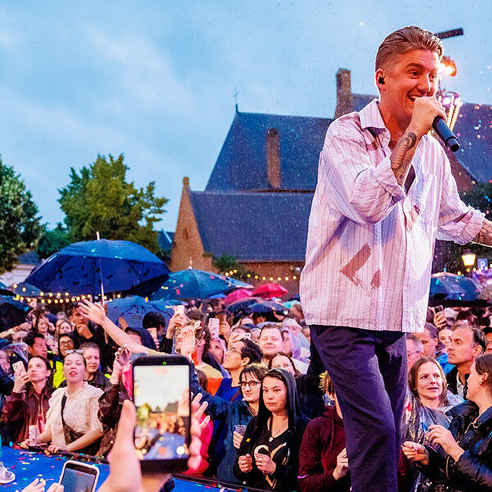 Zanger Mart Hoogkamer op het podium in Moegestel. Op de achtergrond zie je een kerk en heel veel publiek, met paraplu's.