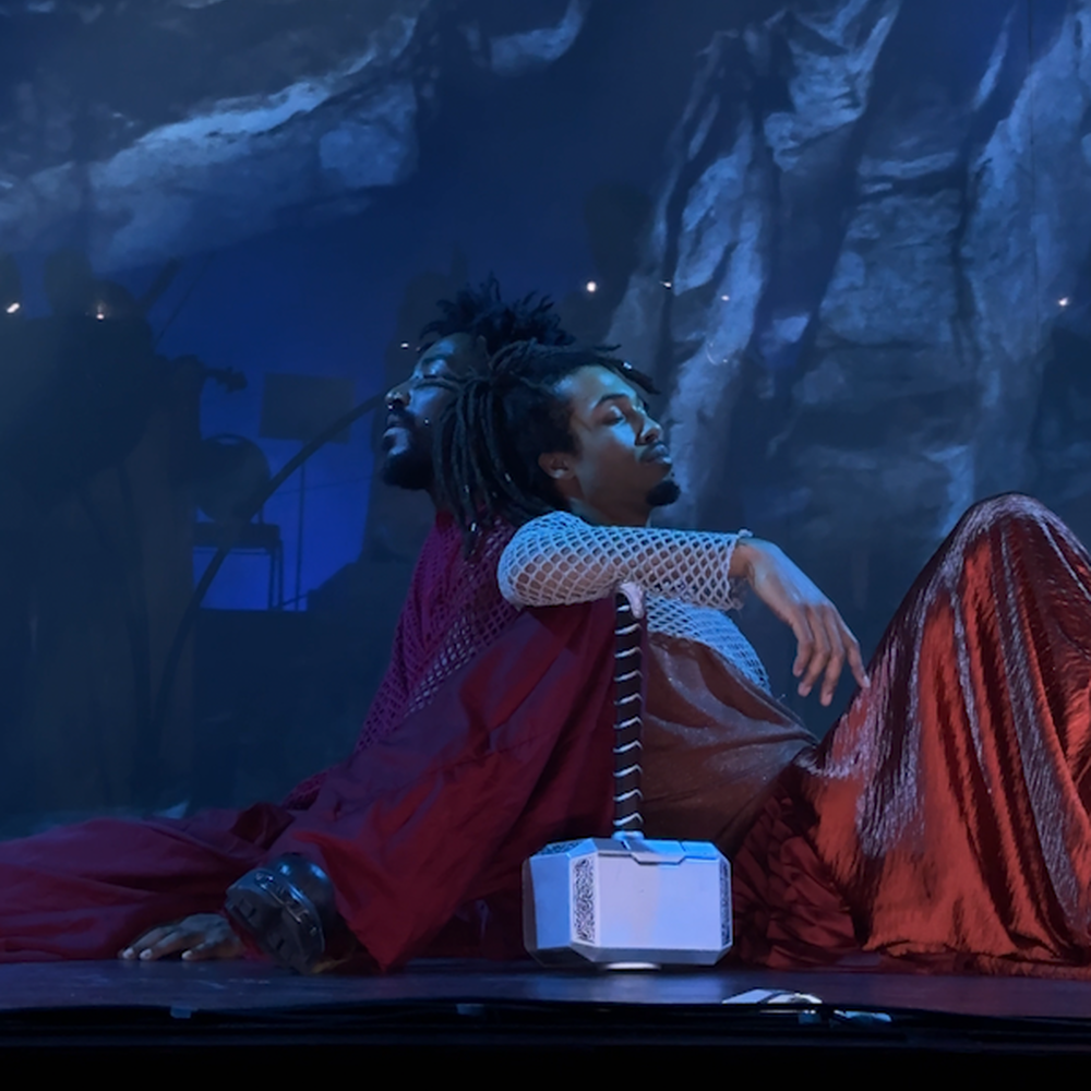 je ziet 2 acteurs van kleur met de grijze ijzeren hamer van Thor, de Noorse god. zij vertolken de rollen van Thor en Loki in oranje en rode gewaden.