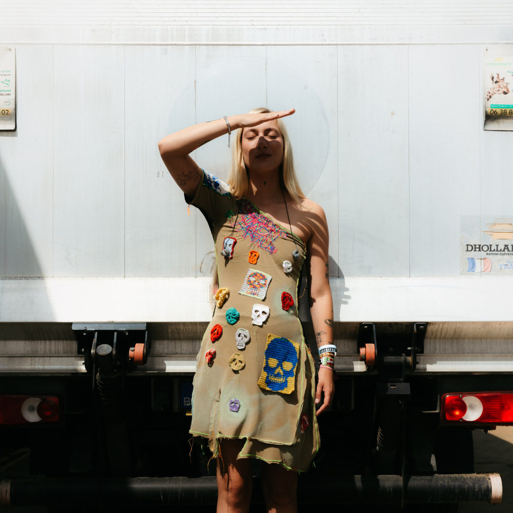 Foto van artiest Stien die voor een vrachtwagen staat en tegen de zon in kijkt. Ze heeft lang blond haar en een jurk vol opgenaaide stukjes stof met doodshoofden erop. De jurk is bruin. Ze houdt haar hand half voor haar gezicht om de zon uit haar ogen te houden en heeft haar ogen dicht.