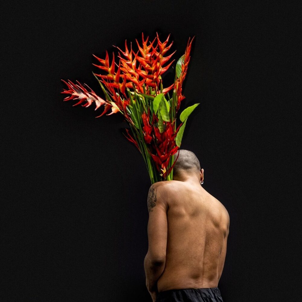 Een man met ontbloot bovenlichaam met zijn rug naar de camera toe. Hij houdt takken met rode bloemen vast die vel afsteken tegen de zwarte achtergrond.