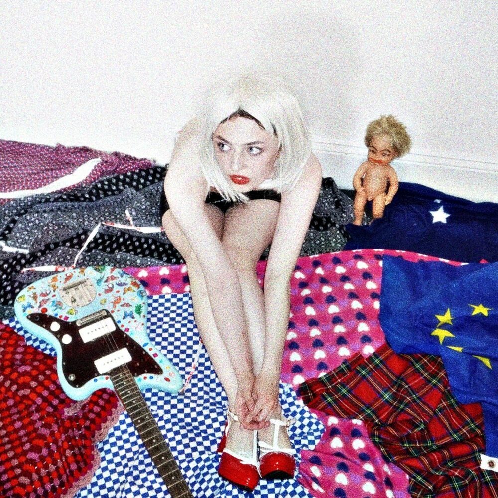 Een foto van de artiest TTSFU. Ze zit op een bed waar veel gekleurde dekens op liggen. Naast haar ligt een lichtblauwe gitaar. Ze heeft licht blond haar en rode schoenen aan.