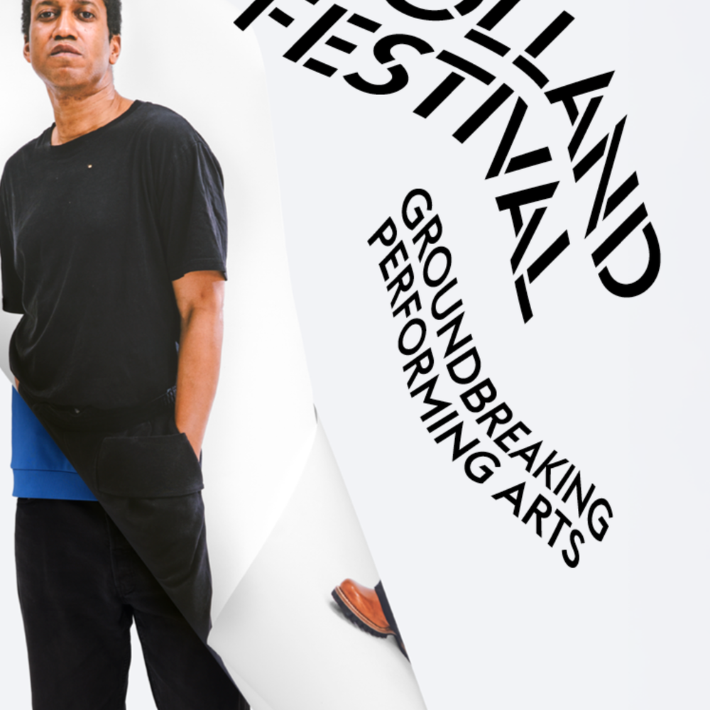 Een foto van Trajal Harell, de choreograaf. Je ziet hem ten voeten uit, met naast hem letters 'holland festival'. Hij staat voor een witte achtergrond en heeft zwarte kleding aan.