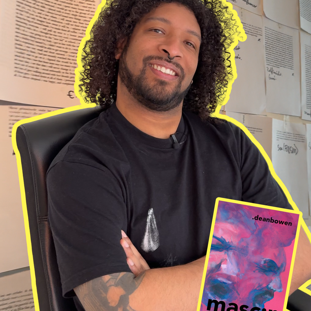 Een foto van Dean Bowen in zijn werkkamer. Hij zit op zijn bureaustoel en draagt zwarte kleren en kijkt vrolijk de camera in. Hij heeft zijn armen over elkaar en een grote bos krullen.