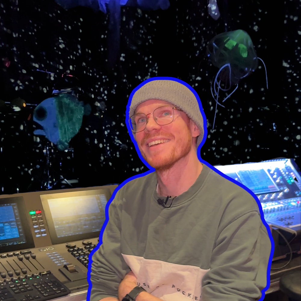 Een foto van Douwe, de lichtontwerper die geïnterviewd wordt over de voorstelling DIEP. Hij kijkt lachend de camera in en zit voor zijn lichttafel met zijn armen over elkaar. Hij heeft een bril en een grijze muts op. Op de achtergrond achter de lichttafel is een soort diepzee te zien, met kleine lichtjes en zeedieren als oplichtende kwallen en neonvissen.