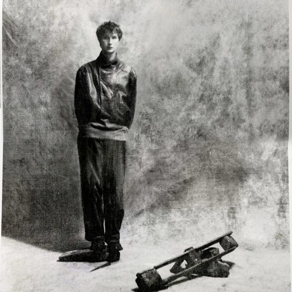 Een foto van artiest Dove Ellis. Het lijkt een hele oude foto door de soort grijze washing. Hij staat voor een grijs studio doek en je ziet hem van top tot teen. Hij draagt donkere kleding en heeft kort haar. Op de grond naast hem ligt een vaag object.