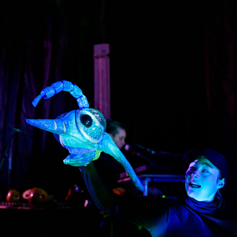 Een vrouw met een pop van een onderwaterdier. Het ziet er futuristisch uit, met neon kleuren en een donkere achtergrond. De vrouw kijkt naar de pop in haar hand.