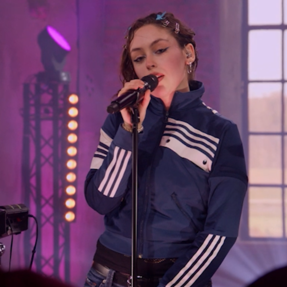 een afbeelding van zangeres IDA die zingt. De setting is een oude fabriekshal en je ziet haar vanaf haar middel. Ze zingt en heeft haar ogen dicht. Ze draagt een donkerblauw vest en de lichten zijn paars.