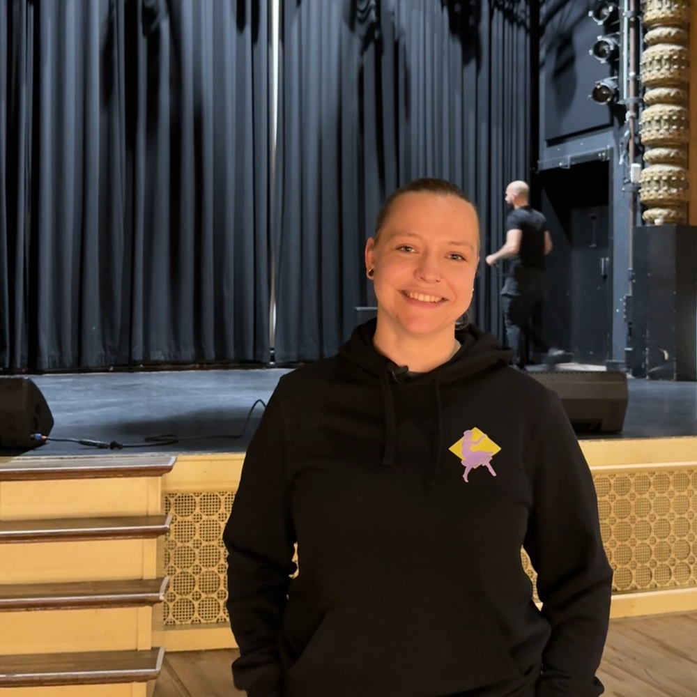 Izanka, de geïnterviewde, in de zaal. Ze heeft zwarte kleren aan en heeft haar handen in haar zakken. Ze kijkt lachend de camera in.