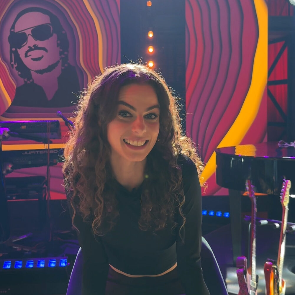 Yvette zit op een stoel in de tv studio van The Best of Pop en kijkt de camera in. Ze heeft lange bruine krullen en lacht.