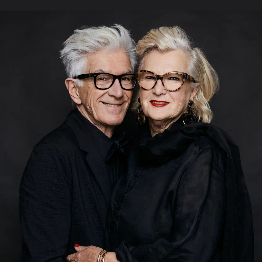 Een foto van Puck en Hans een paar jaar geleden. Ze staan in een fotostudio voor een donkere achtergrond en houden elkaar liefdevol vast. Ze dragen allebei een bril. Hans heeft grijs haar en Puck grijs-geblondeerd haar en rode lippenstift op. Ze hebben allebei zwarte kleren aan.