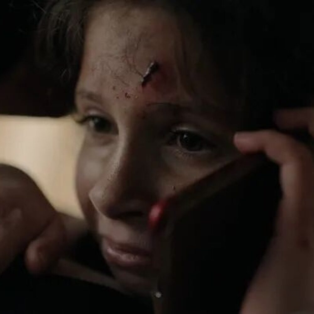 je ziet een jong meisje met een wond op haar hoofd. Ze is aan het bellen met een angstige blik.