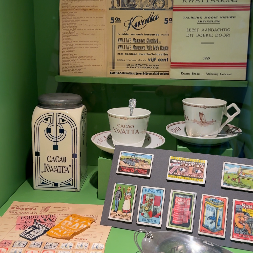 Een foto van de vitrine met Kwatta chocolade. Het is een groene kast, met een blik waar 'cacao Kwatta' opstaat, kopjes met dezelfde tekst en verschillende krantenknipsels en plaatjes met grafische ontwerpen van Kwatta.