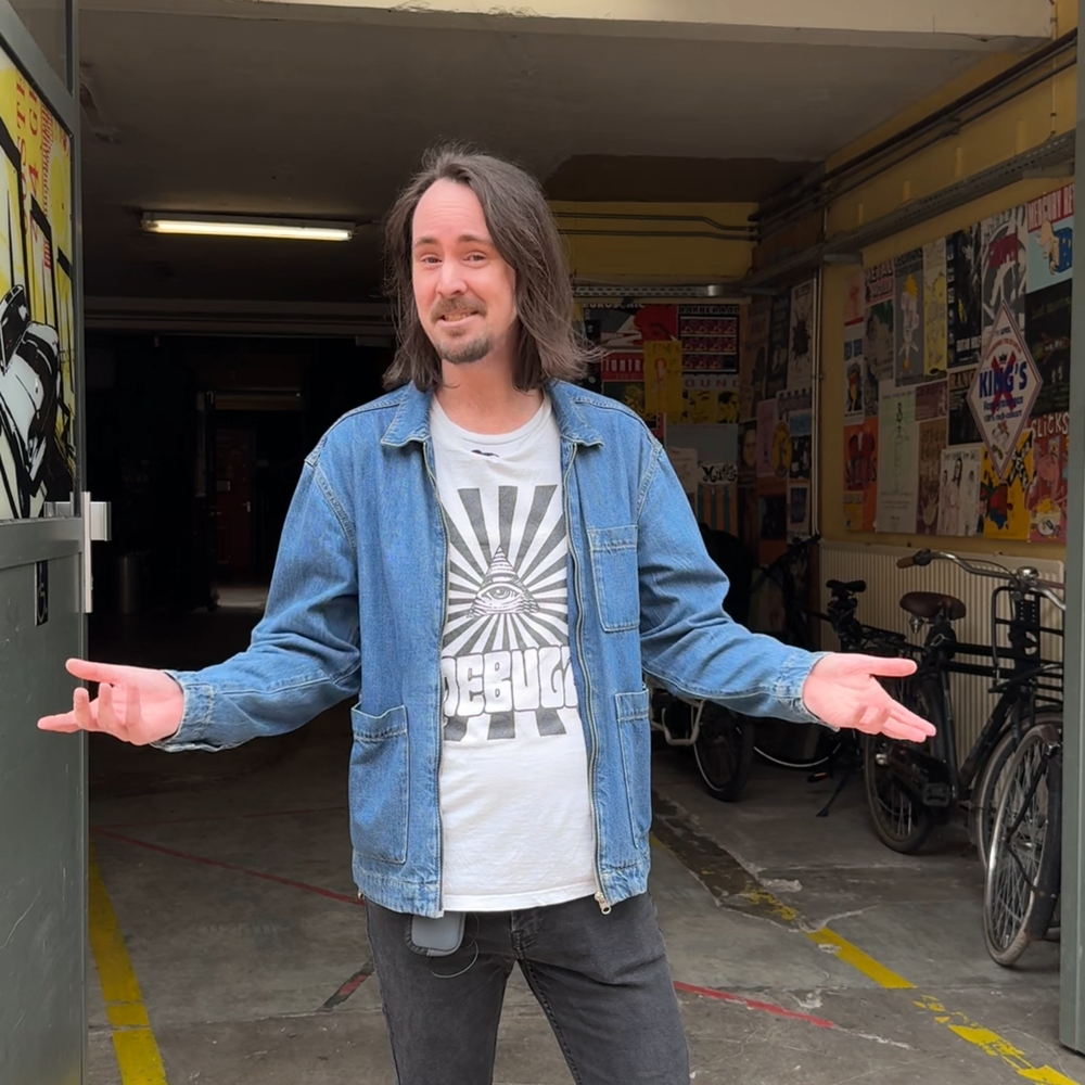 Artiestenbegeleider Mark in de deuropening van de garage van VERA. Hij heeft beide armen naar buiten gedraaid, een open houding, en kijkt vriendelijk naar de camera. Alsof hij je verwelkomd in via de artiesteningang van VERA.