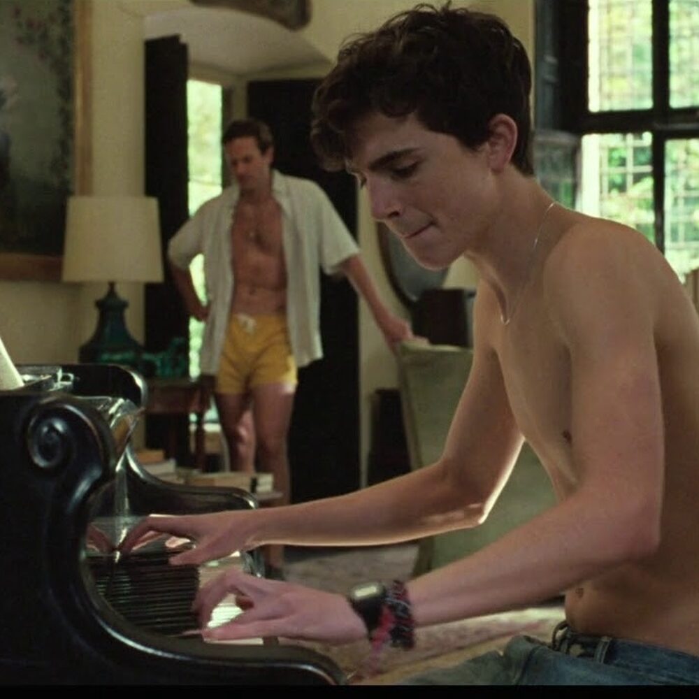 Elio speelt piano in de woonkamer terwijl Oliver staat te luisteren.