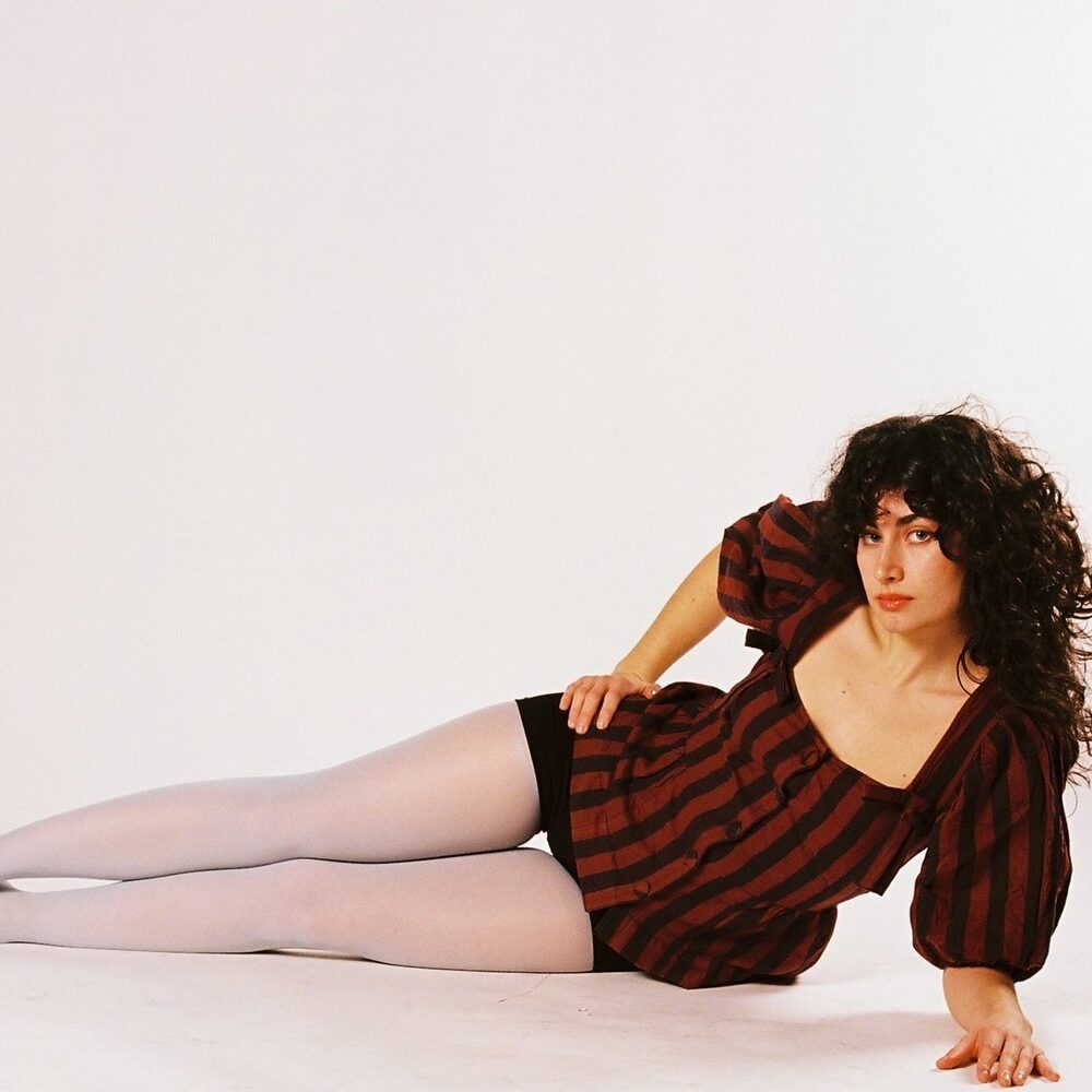 Een foto van de artiest Min Taka. Ze ligt in een witte studio met haar benen links op de foto en haar lijf naar rechts. Ze steunt op een arm en heeft de andere arm in haar zij. Ze heeft een kort rood jurkje aan met zwarte strepen en losse zwarte krullen. Ze kijkt de camera in.