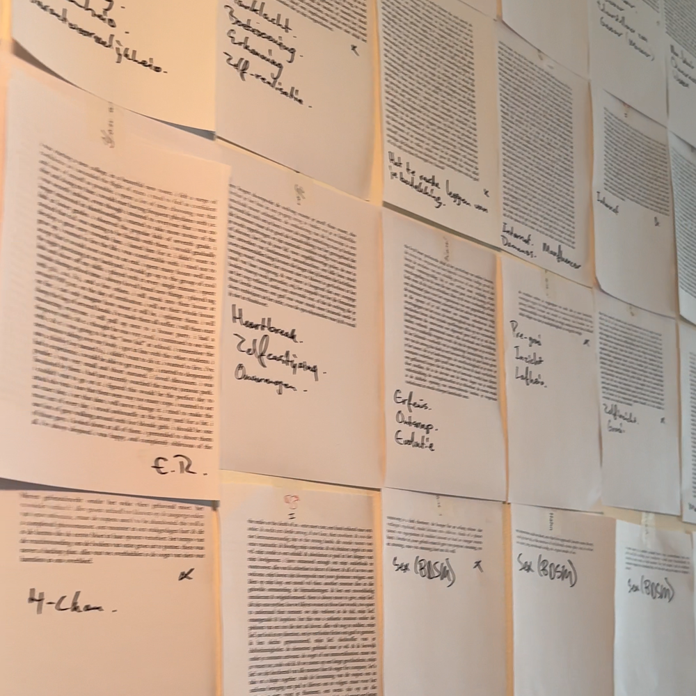 Een foto van de gedichten op de muur. Het is een witte muur met allemaal uitgeprinte blaadjes met nog met de hand geschreven notities erdoorheen opgeplakt. Heel veel losse blaadjes op een muur.