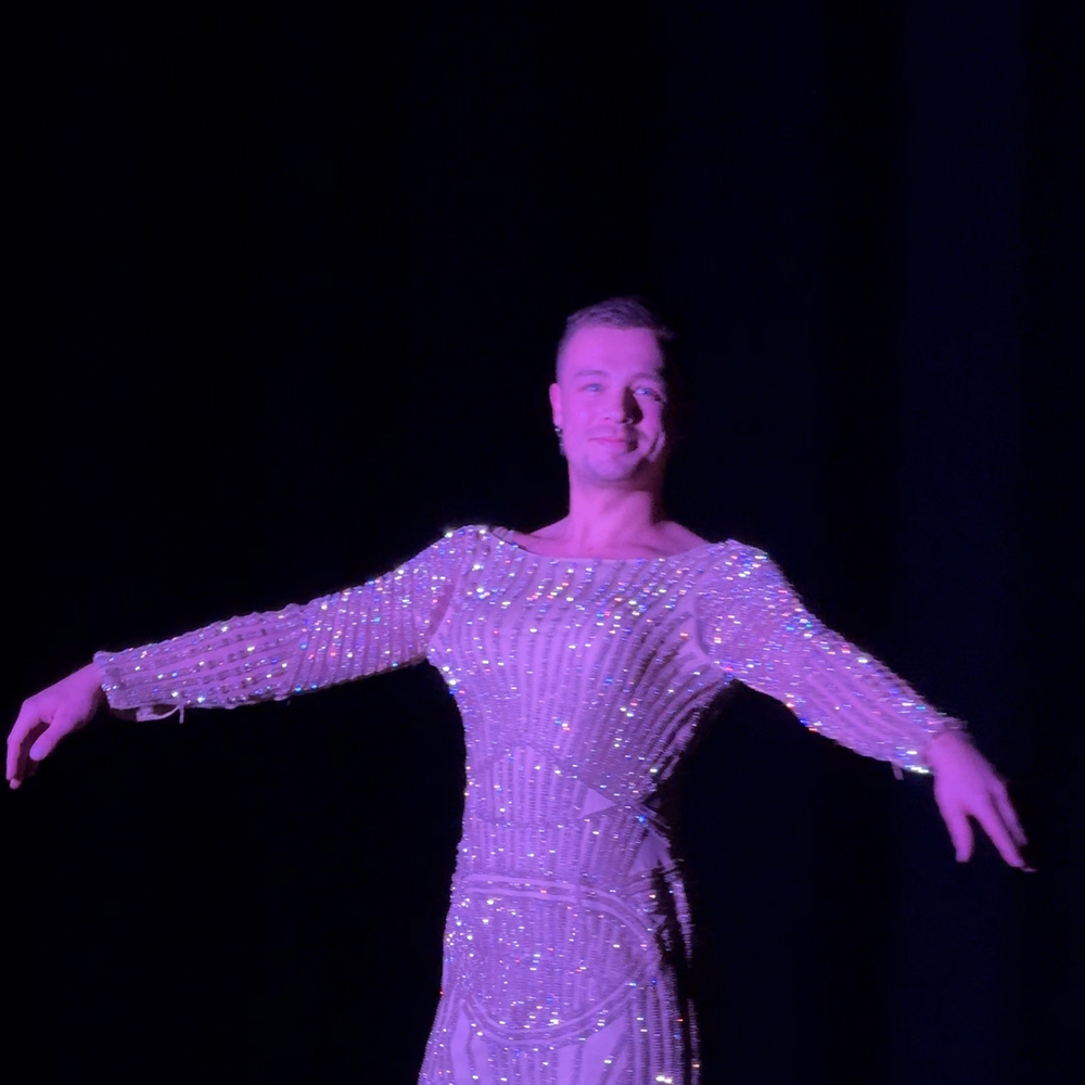 Een stilbeeld van Nick uit de voorstelling. Hij draagt een super glitterende gala jurk en kijkt trots richting de camera.