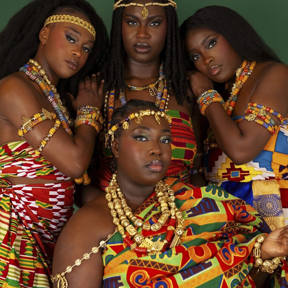 Een foto van vier zwarte vrouwen in ontzettend kleurrijke kleding. Ze kijken alle vier strak naar de camera en leunen op elkaar.
