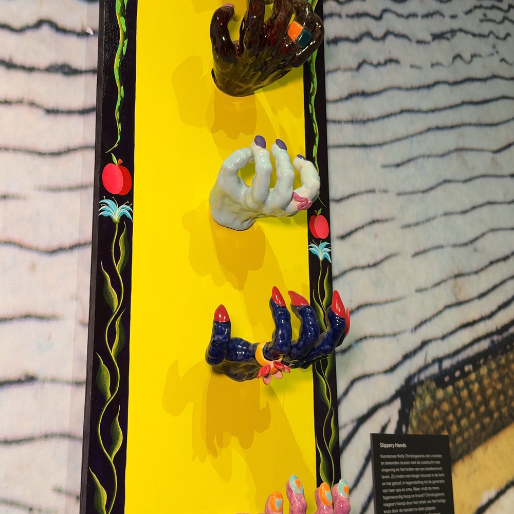 Een foto van het kunstwerk Slippery Hands. Het is een langgerekt kunstwerk wat aan de muur haght. Tegen een velgele achtergrond hangen meerdere, gekleurde handen onder elkaar die allemaal iets vast lijken te grijpen. Als je goed kijkt, zie je dat er een visdraad door de handen van boven naar beneden loopt, waar een klein soort splintertje van klei aanhangt.