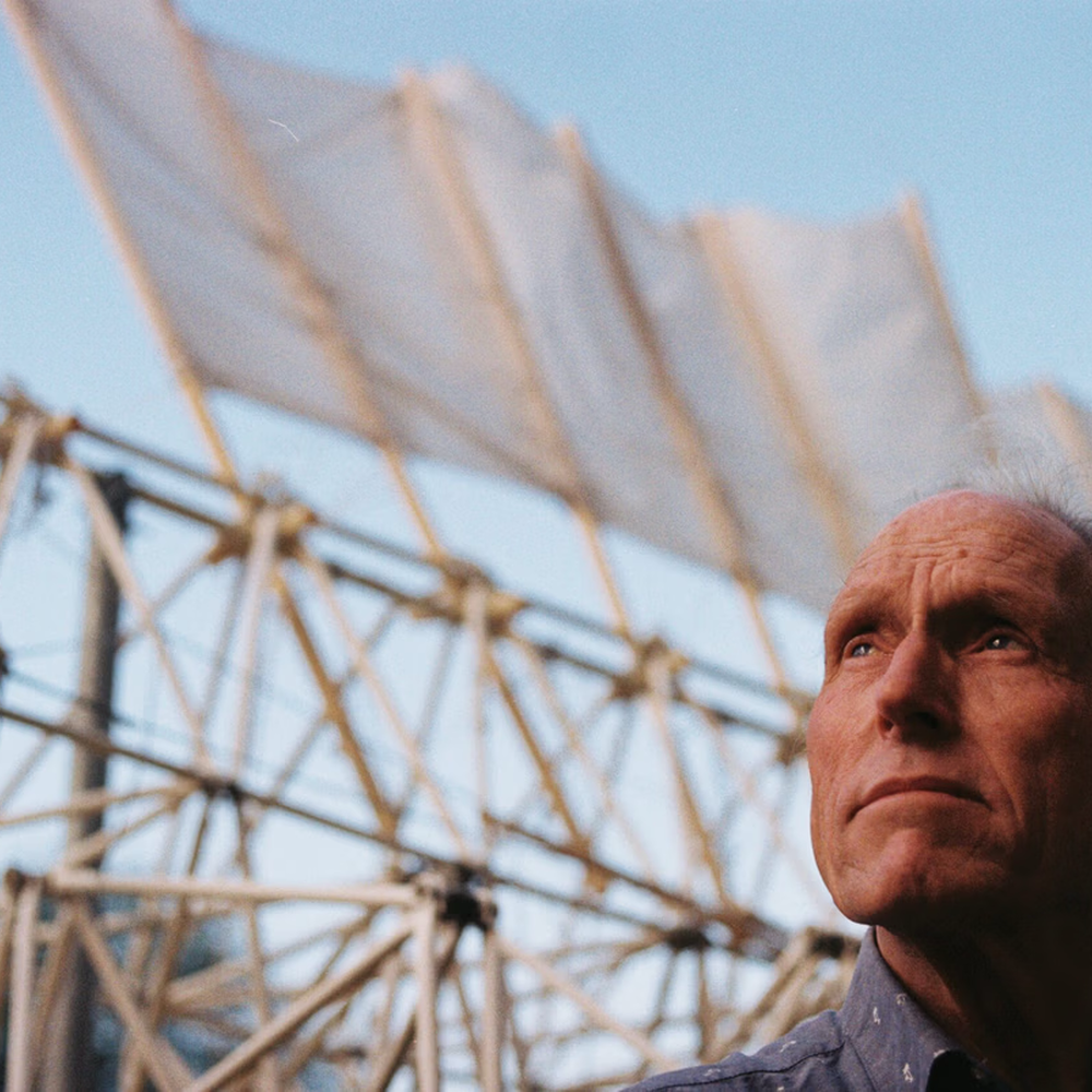 Een foto van de kunstenaar die rechts onderin beeld trots naar linksboven kijkt. Achter hem zie je een van zijn strandbeelden. Het is een soort posterbeeld wat bij de docu hoort.