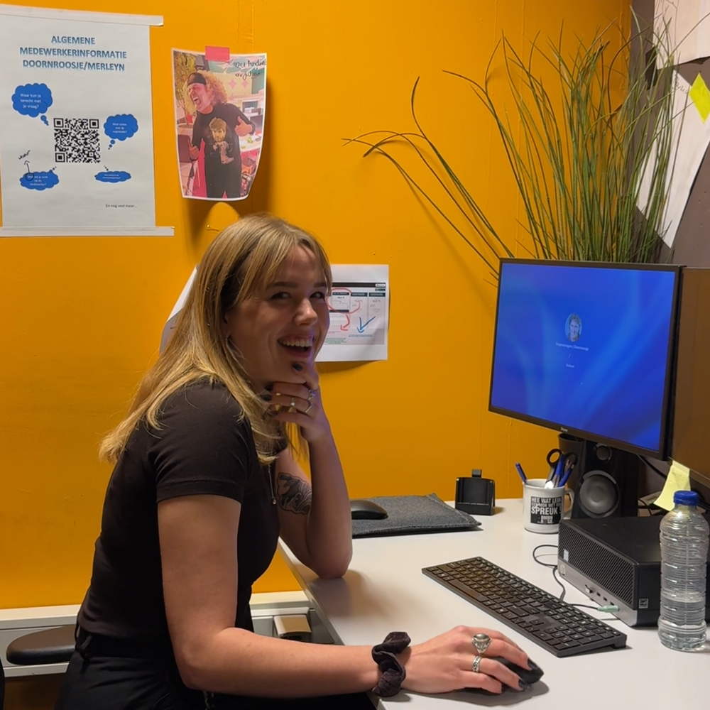Susanne achter de computer. Ze kijkt lachend naar de camera. De muur is geel en er staat een plant op het bureau.