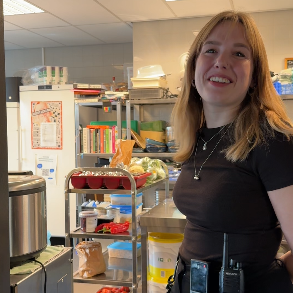 Een foto van Susanne die bij een keuken staat. Susanne is een jonge meid met los blond haar en ze kijkt lachend naar de camera. Achter haar zie je een grote professionele keuken.