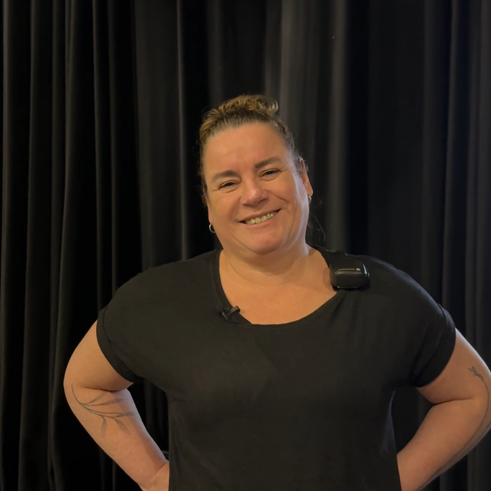 Ilja, die de vragen in dit stuk beantwoordt. Ze staat met haar handen in haar zij en lacht naar de camera. Ze heeft zwarte kleren aan en staat voor een zwart gordijn.