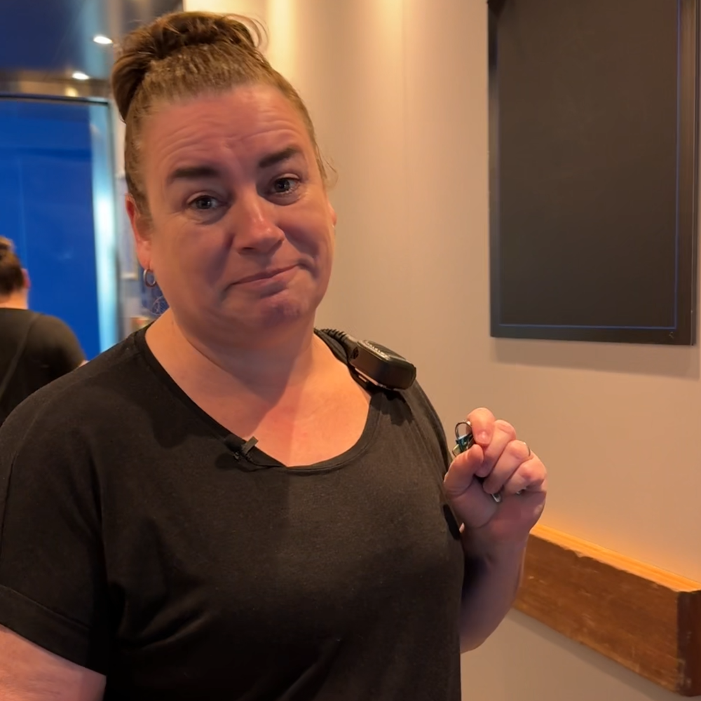 Een foto van Ilja, die de vragen in de tekst beantwoordt. Ze kijkt vriendelijk de camera in. Ze staat ergens in een gang en heeft zwarte kleren aan en een walkietalkie op haar schouder.