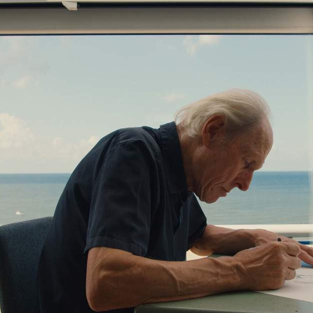 Een foto van de kunstenaar die achter een tafeltje zit met een vel papier. Hij zit voorover gebogen en is aan het tekenen. We zien hem van de zijkant. Op de achtergrond zie je de zee en het strand.