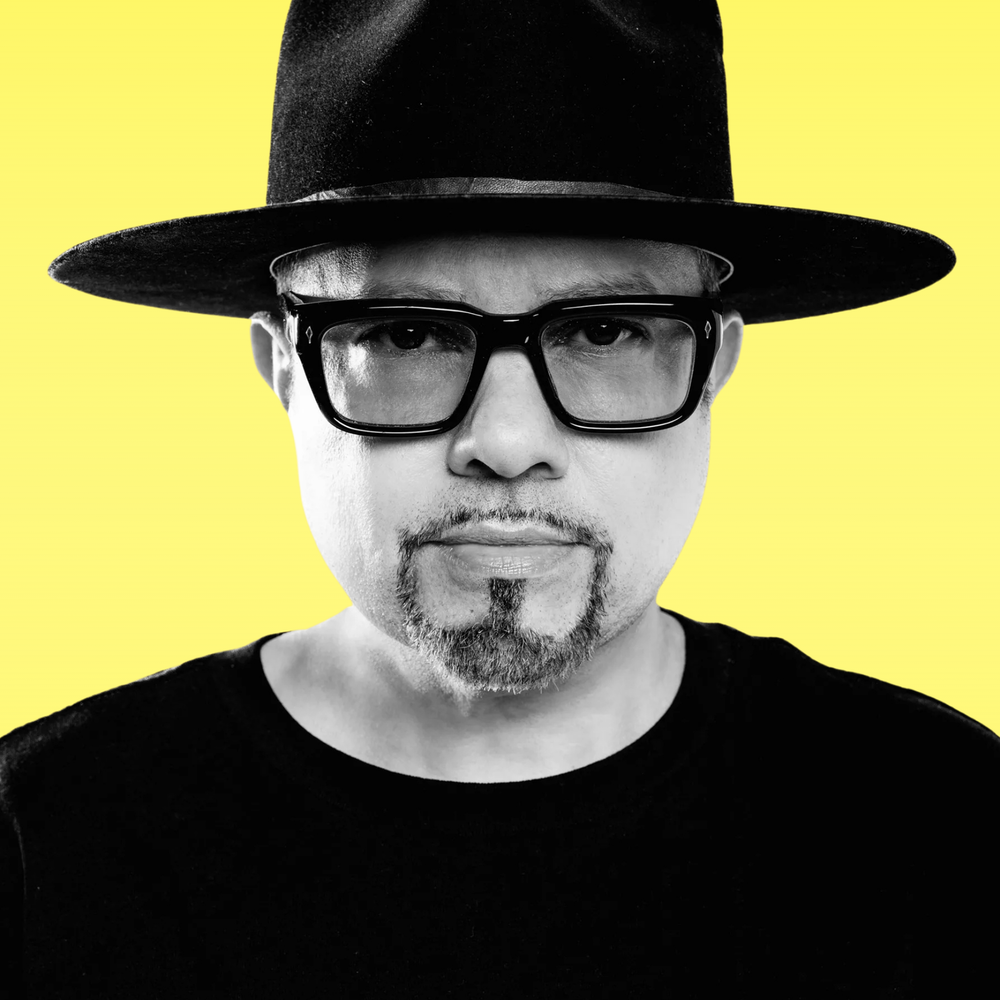 Foto van Louie Vega. Een man met een hoed, bril en baard en snor, helemaal in zwarte kleren voor een gele achtergrond.