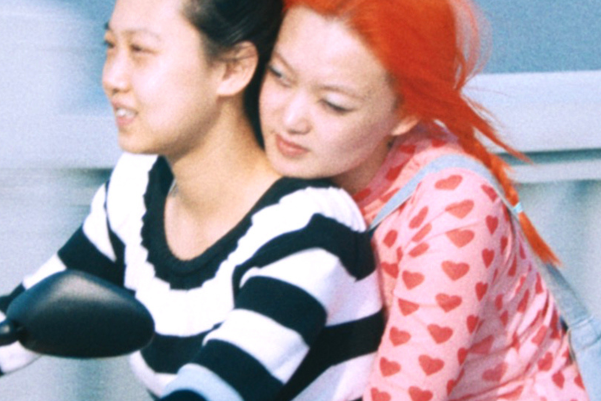 Twee Chinese actrices uit de film 1 Girl Infinite zitten samen op een scooter. Degene die rijdt, draagt een zwart/wit gestreept shirt en heeft zwart haar. Degene die achterop zit, houdt de andere persoon vast om haar middel. Ze draagt een roze shirt met rode hartjes, een groen rokje en heeft felrood haar.