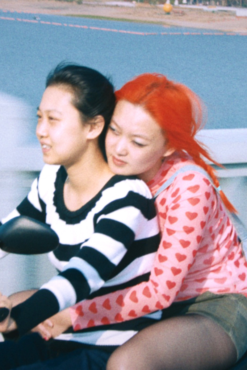 Twee Chinese actrices uit de film 1 Girl Infinite zitten samen op een scooter. Degene die rijdt, draagt een zwart/wit gestreept shirt en heeft zwart haar. Degene die achterop zit, houdt de andere persoon vast om haar middel. Ze draagt een roze shirt met rode hartjes, een groen rokje en heeft felrood haar.