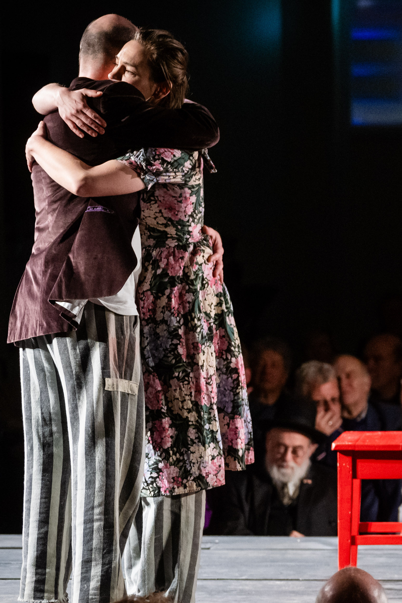 Op de foto zijn twee knuffelende personen op een podium te zien. Een persoon, de vrouw, heeft een bloemetjesjurk aan tot over haar knieën. Daaronder draait ze een loszittende grijs-gestreepte broek. De man draagt een bruin colbert. Ook hij draait dezelfde soort grijs-gestreepte broek. Naast hen op het podium staan een rode tafel en stoel.