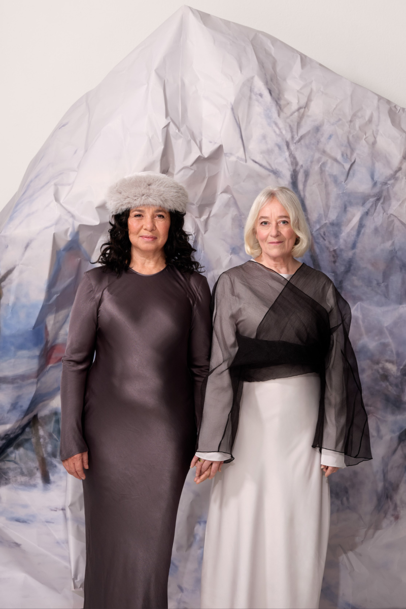 Twee vrouwen staan naast elkaar met hun vingers in elkaar verweven. Achter hen staat een ijsberg van papier. De linker vrouw heeft donkerbruin krullend haar, draagt een grijze muts en een lange bruine jurk. De rechter vrouw heeft kort, stijl en blond haar en draagt een lange zwart/witte jurk.