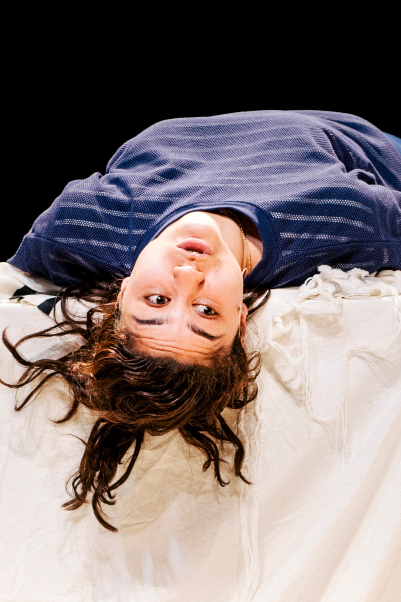 Actrice Soumaya ligt in blauwe kleding met haar rug op een tafel met een groot wit doek. Haar hoofd hangt over de tafel heen en ze kijkt naar rechts.