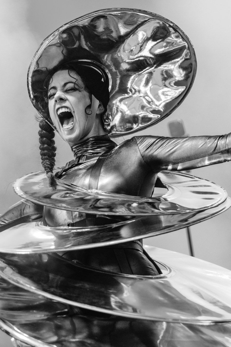 Zwartwitfoto van een vrouw die met haar armen wijd op een podium staat. Haar mond heeft ze wijd open en ze heeft een alienachtig kostuum aan dat bestaat uit een soort schotels. Op haar hoofd draagt ze ook een schotel.