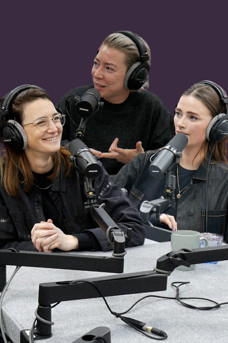 Drie jonge witte vrouwen zitten aan een grijze tafel met microfoons. Ze hebben allemaal een zwarte koptelefoon op. De linker vrouw heeft middelkort bruin haar. Ze draagt een lichte ronde bril en een zwarte blouse. Op haar gezicht heeft ze een glimlach. De middelste vrouw heeft kort blond haar en draagt een zwarte trui. Ze kijkt lachend naar links. De rechter vrouw heeft lang bruin haar en draagt een overhemd van grijze en donkerblauwe spijkerstof. Ze kijkt neutraal naar links.