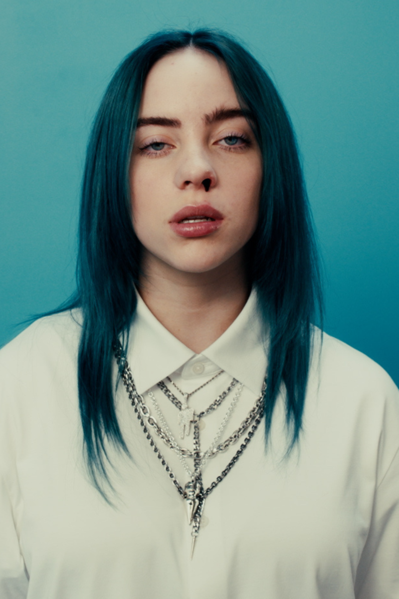 Zangeres Billie Eilish staat voor een blauwe muur. Ze heeft blauw haar, draagt een wit overhemd met kettingen erover en er komt bloed uit haar neus.