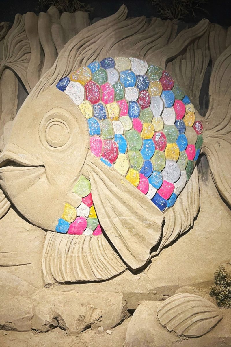 Zandsculptuur van de vis uit het kinderboek 'De mooiste vis van de zee'. Zijn kop en vinnen zijn zandkleurig (beige) en zijn schubben zijn alle kleuren van de regenboog (blauw, roze, groen, rood, wit, geel).