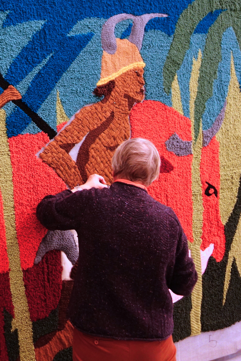 Er wordt door twee vrouwen gewerkt aan het wandtapijt. Op het tapijt staan twee personen, gebroken ketens, nootmuskaat en een palmboom.