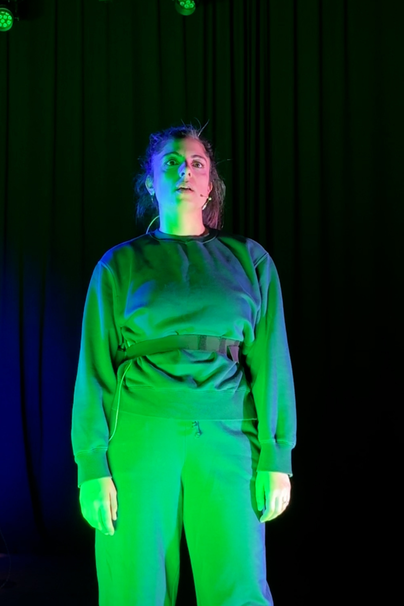 Een actie foto van Yeliz op het toneel. Ze heeft een blauw pak aan en staat in fel groen licht. Ze kijkt serieus en vlak langs de camera.