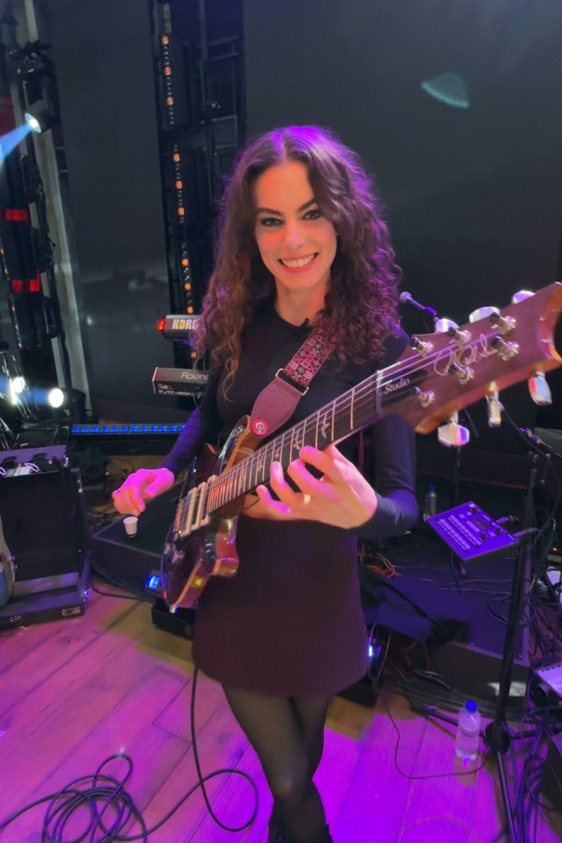 We zien Yvette met haar gitaar in haar handen. Ze staat in een tv studio en kijkt de camera in en lacht. Ze speelt op haar gitaar.