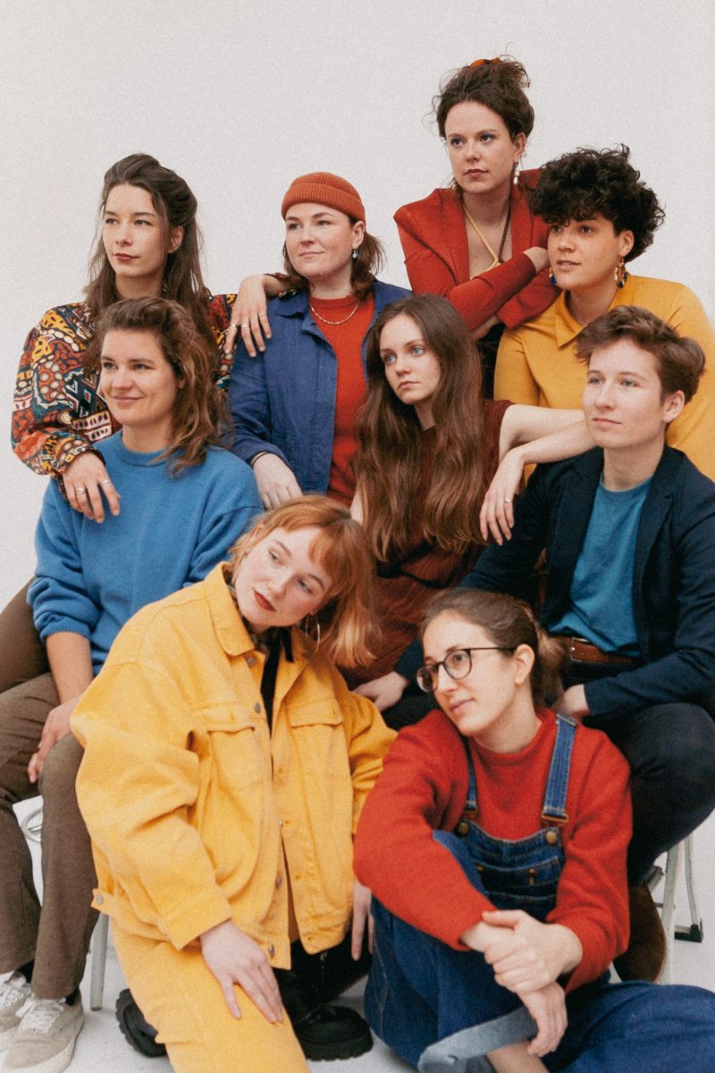 Dit is de persfoto van PARRA.DICE met de negen vrouwelijke/non-binaire bandleden. Ze zijn gekleed in het kleurpalette rood, blauw en geel.