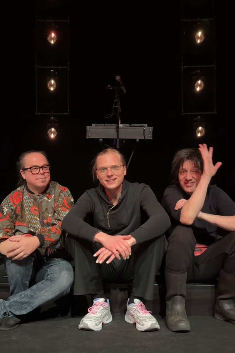 De drie leden van de band Prins S en de Geit zitten op een klein podiumpje en kijken naar de camera. Ze lachen allemaal vriendelijk en de meest rechter zwaait.