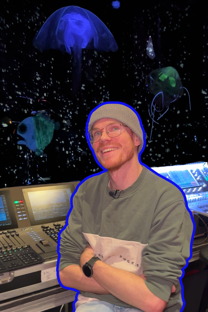 Een foto van Douwe, de lichtontwerper die geïnterviewd wordt over de voorstelling DIEP. Hij kijkt lachend de camera in en zit voor zijn lichttafel met zijn armen over elkaar. Hij heeft een bril en een grijze muts op. Op de achtergrond achter de lichttafel is een soort diepzee te zien, met kleine lichtjes en zeedieren als oplichtende kwallen en neonvissen.