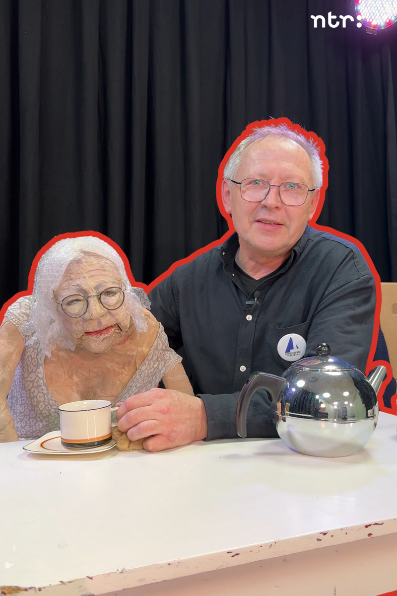 Een foto van Servaes de poppenspeler en de pop die zijn moeder voorstelt. Ze zitten samen achter een witte tafel en er staan een theepot en kopjes. Servaes is een man met een bril en grijs haar en hij draagt een zwart overhemd en kijkt vriendelijk de camera in. De pop naast hem is een oude vrouw, gemaakt van proppen papier en tape, met een bleke huid, bril, bloemenjurk en haar van bubbeltjesplastic.