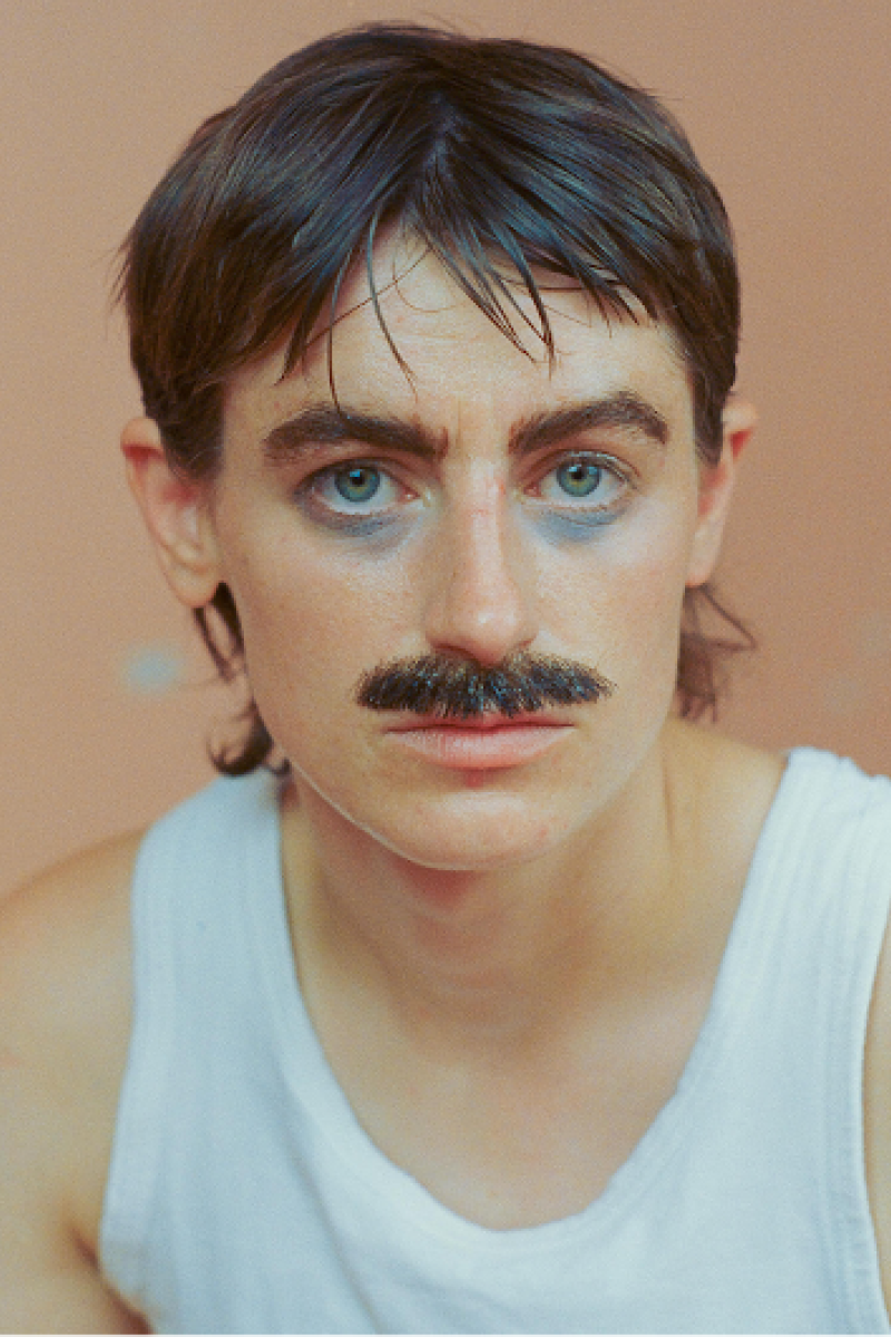 Een foto van Elmer. Ze zit tegen een licht oranje achtergrond en kijkt recht de camera in. Ze heeft een snor en een wit hemd aan.