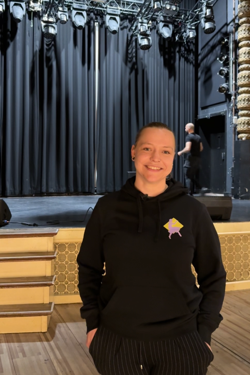 Izanka, de geïnterviewde, in de zaal. Ze heeft zwarte kleren aan en heeft haar handen in haar zakken. Ze kijkt lachend de camera in.