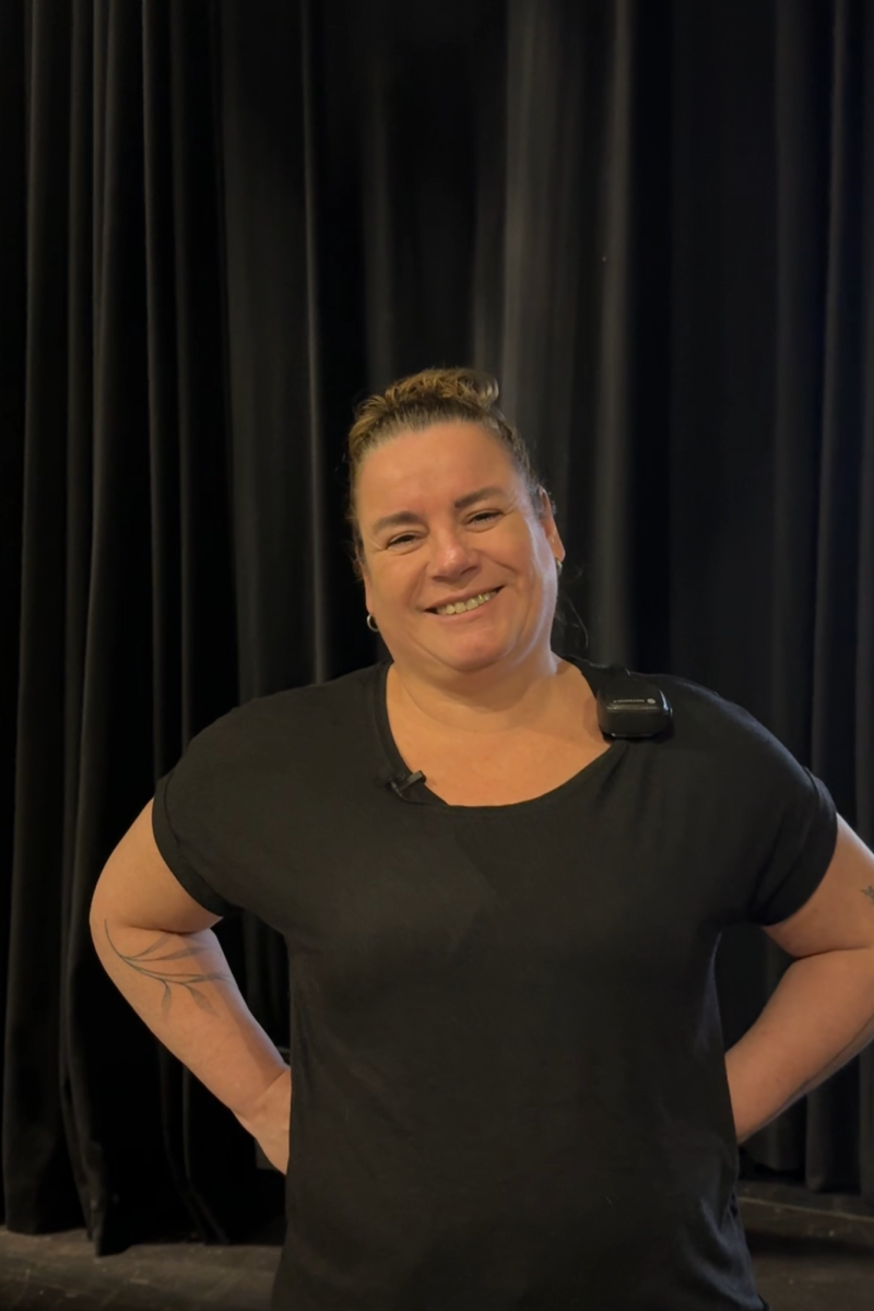 Ilja, die de vragen in dit stuk beantwoordt. Ze staat met haar handen in haar zij en lacht naar de camera. Ze heeft zwarte kleren aan en staat voor een zwart gordijn.