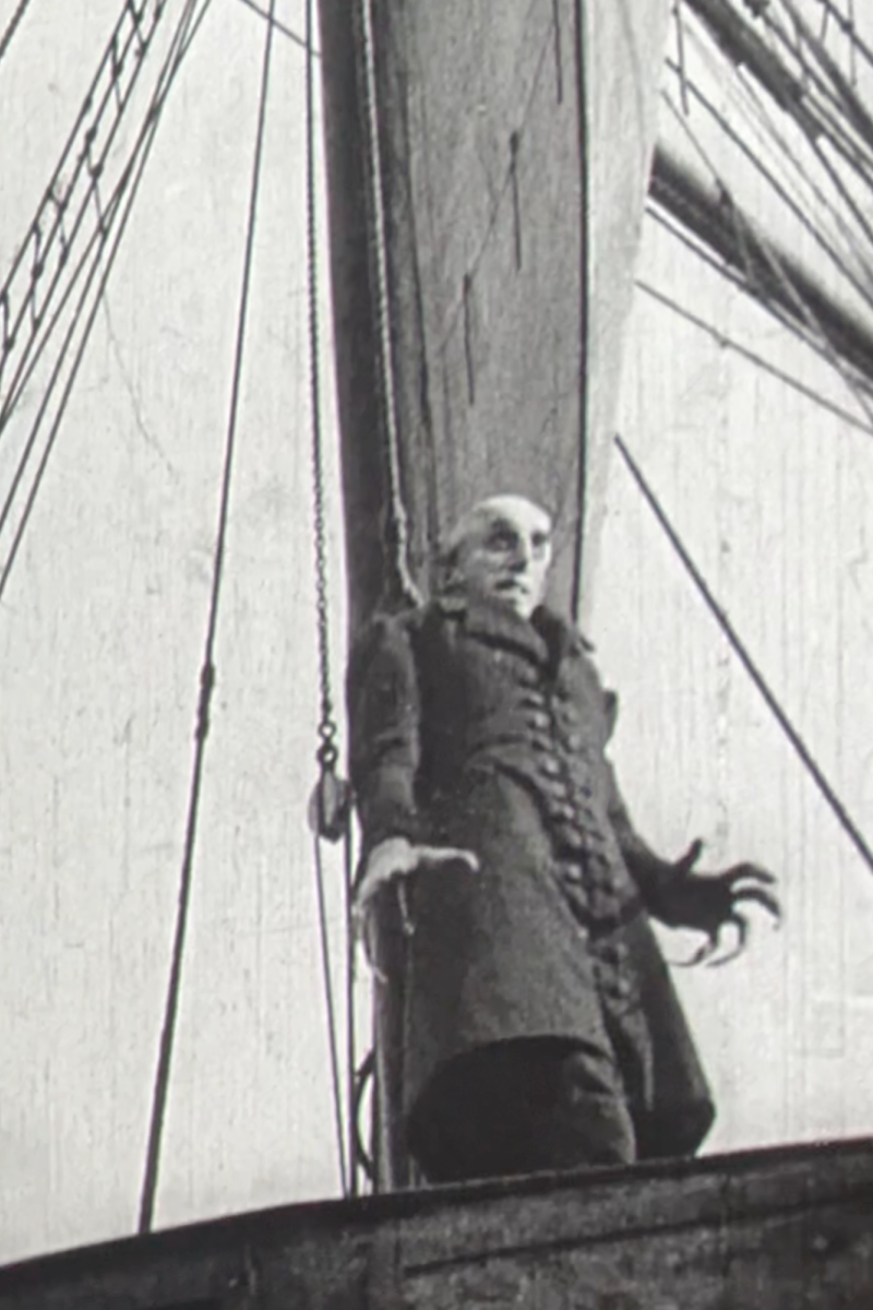 Een stil uit de film Nosferatu. We zien een griezelige man op een schip staan. Hij is erg lang en heeft hele lange vingers.